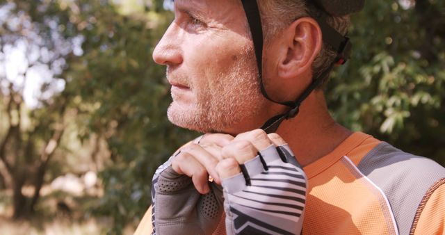 A senior man in athletic clothing is preparing for a cycling session, adjusting his helmet while wearing cycling gloves. He is outdoors, in a wooded area with bright sunlight. This image can be used for promoting senior fitness, outdoor activities, or cycling safety tips.