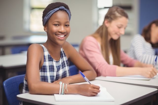 Young African American girl smiling while writing in a classroom. Ideal for educational content, school brochures, academic websites, and learning-related advertisements.