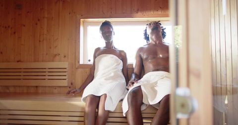 African american couple relaxing and holding hands in wooden sauna wrapped in white towels