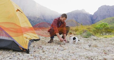 Crouching camper driving tent stake on rocky mountain plain with curious dog observing