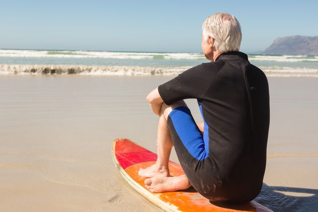 Rear view of senior man sitting on surfboard against sea at beach