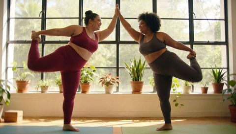 Curvy women practicing partner dancer pose in sunlit yoga studio with indoor plants