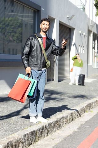 Young man holding smartphone and shopping bags on urban curb in leather jacket streetstyle
