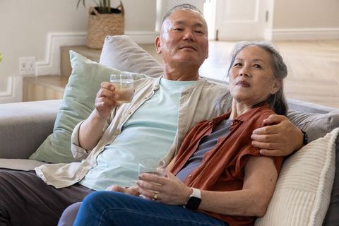Asian senior couple relaxing on sofa holding glasses, enjoying cozy home companionship