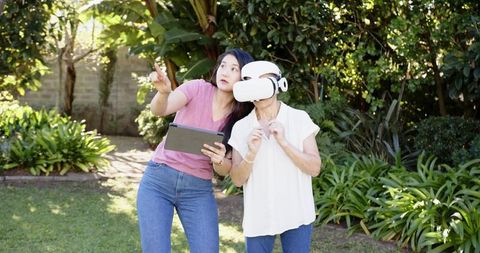 Asian women guiding older woman through virtual reality in sunlit garden with tablet