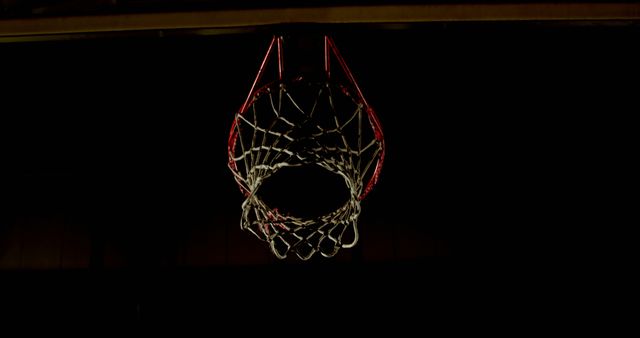 This stock image captures a basketball hoop in dim lighting against a dark background. The focus on the hoop highlights the importance of the scoring element in basketball. Ideal for articles, advertisements, or promotions related to sports, athletics, fitness, basketball training, teamwork, motivation, or competitions.
