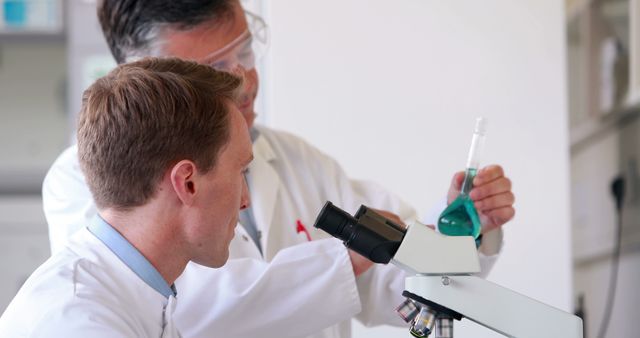 Researchers in lab coats working together in a modern laboratory with scientific equipment. One scientist is focusing through a microscope while the other is holding a test tube with green liquid. Ideal for content related to scientific research, medical technologies, laboratory work, biomedical advancements, and teamwork in science. Use this for articles, presentations, or educational materials to convey collaboration and innovation in science.