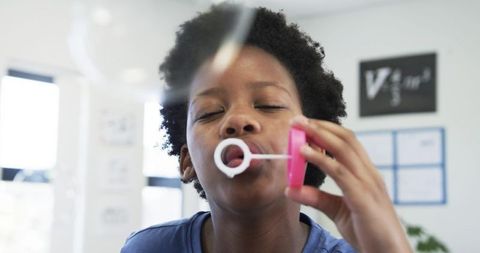African girl blowing soap bubbles in bright classroom holding pink wand and jar