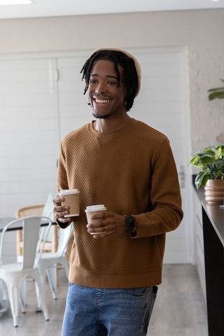 Smiling african american man holding two takeaway coffees in sunlit cafe wearing quilted sweater