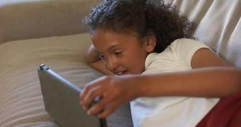 Young girl reclining on beige sofa holding tablet in protective case watching video