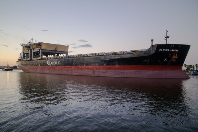 Image showing a large cargo ship anchored in a calm harbor during sunset. The sky is slightly cloudy and reflects on the tranquil water, creating a peaceful atmosphere. Ideal for use in articles about maritime industry, global shipping, and cargo transport, as well as promotional materials for shipping companies or marine logistics.