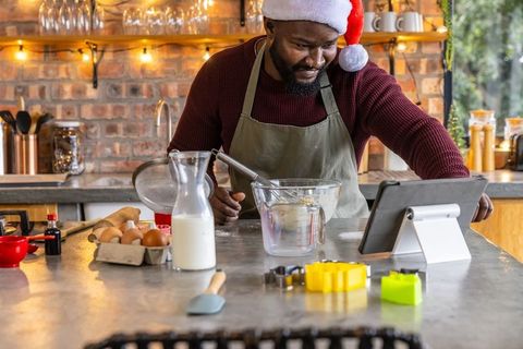 African american man wearing santa hat mixing cookie batter and using tablet in kitchen