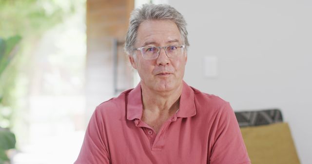 Senior man wearing glasses and a red shirt, sitting indoors and looking confident. This can be used for promotional materials related to senior living, healthy aging, and personal stories of elderly life. Perfect for articles about retirees, blogs on elderly lifestyle, or advertisements for senior products.