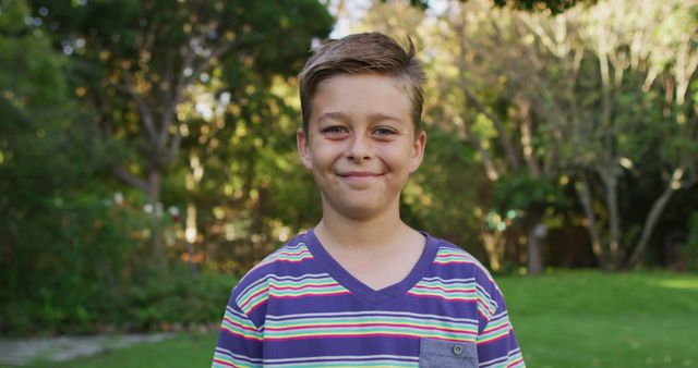 A young boy, wearing a colorful striped shirt, smiles while standing in an outdoor park filled with lush green trees and grass. The photo conveys a sense of joy, carefree youth, and a connection to nature. Ideal for use in magazines, advertising related to children's fashion or outdoor activities, educational materials, and websites focused on family and youth.
