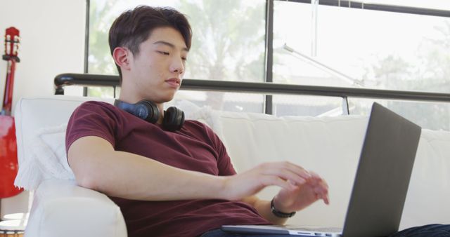 Asian male teenager wearing headphones and using laptop in living room. spending time alone at home.