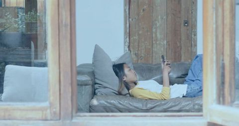 Relaxing asian woman lying on sofa scrolling phone through wooden window frame in cozy living room