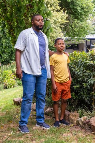 African american father posing with son in backyard garden, smiling with hand on shoulder