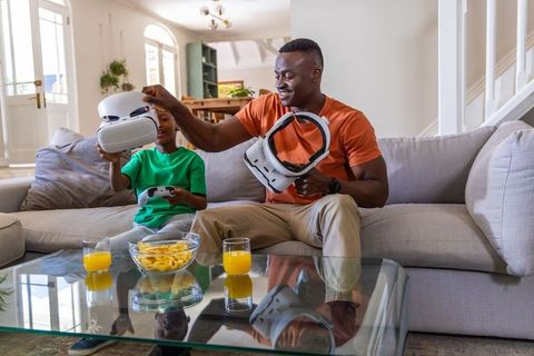 African american father and son sharing vr headsets on couch sunlit living room bonding