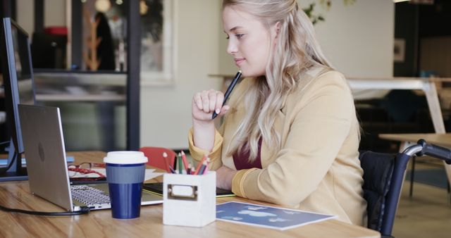 Young woman in wheelchair working at modern office desk with laptop, coffee cup, and documents. She holds a pen and looks focused, emphasizing accessibility and inclusion in professional settings. Ideal for illustrating themes of remote work, accessibility in the workplace, and professional productivity in a diverse work environment.