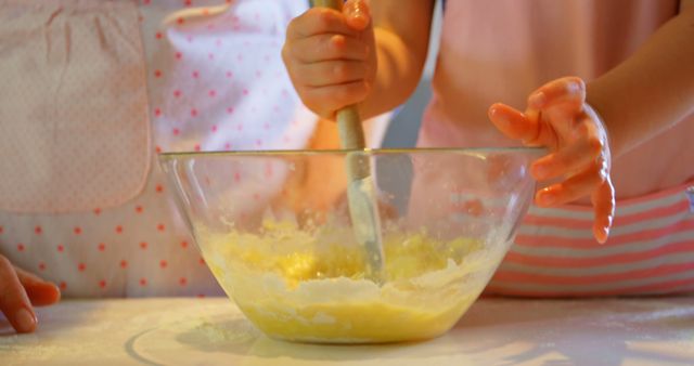 Two children are seen focusing on mixing ingredients in a glass bowl. One child stirs the mixture while the other helps stabilize the bowl. This photo can be used for themes related to family time, cooking activities, children's hobbies, and educational material on baking. Ideal for articles, advertisements, and blogs on fun family activities, cooking tutorials, and childhood development.