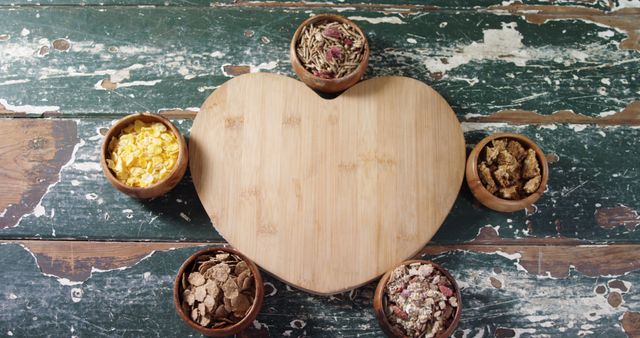 Assortment of nutritious breakfast cereals in wooden bowls arranged around heart-shaped cutting board on rustic wooden table. Ideal for use in food blogs, websites promoting healthy eating, socially-conscious diets, or farm-to-table movements.