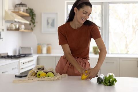 Mid-adult woman preparing fresh fruit with reusable bag in bright modern kitchen