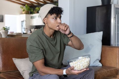 Young african american man watching movie eating popcorn on cozy living room sofa, wearing beanie