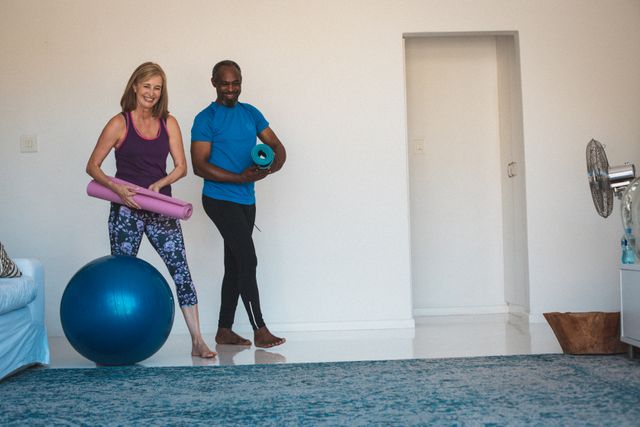 Senior couple smiling and preparing for a yoga session at home. They are holding yoga mats and a stability ball, indicating a focus on fitness and wellness. Ideal for use in articles or advertisements about home workouts, senior fitness, healthy lifestyles, and staying active during quarantine.