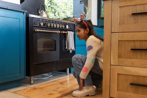 Curious child crouching beside range wearing festive headband in bright kitchen