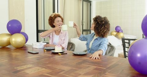 African American women celebrating home birthday party with cake balloons clinking mugs