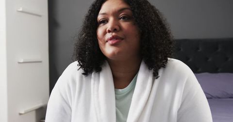 Middle-aged woman wearing white bathrobe sitting on bed calm contemplative intimate bedroom portrait
