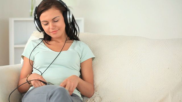 Young woman with long brunette hair sitting on a white sofa in a living room, enjoying music through large headphones. She is casually dressed in a light green t-shirt and grey pants, appearing relaxed and content. The setting suggests a comfortable home environment, perfect for moments of leisure and self-care. Useful for articles or advertisements about relaxation, self-care, music enjoyment, and home lifestyle.