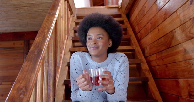 Happy african american woman with afro sitting on stairs and holding cup of tea at home. Lifestyle, free time and domestic life, unaltered.