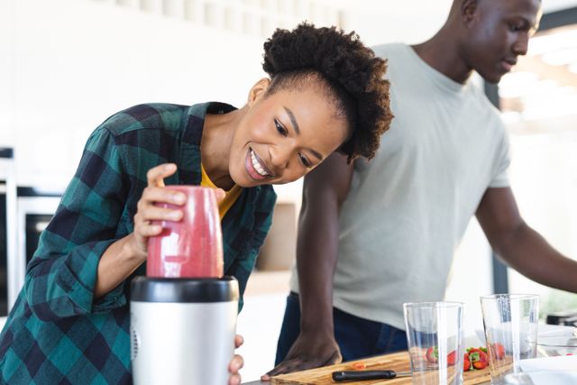 This image shows a joyful African American couple preparing smoothies together in a modern kitchen. The woman is blending ingredients while the man is preparing fruits. Perfect for use in articles or advertisements about healthy living, relationships, home life, or cooking tutorials.