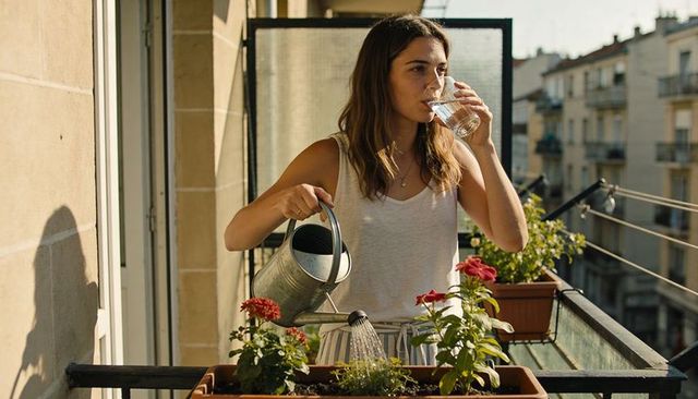 Woman watering balcony plants while sipping water in sunny urban apartment