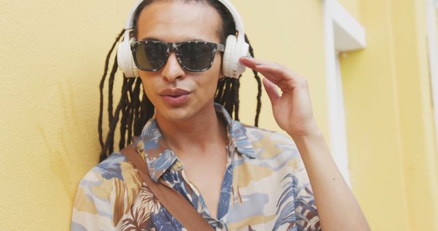 Biracial young professional wearing headphones, standing by yellow wall. He has dreadlocks, light brown skin, and is wearing patterned shirt
