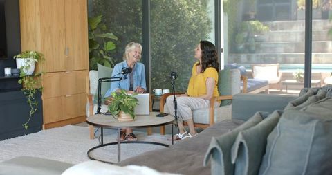 Senior woman and hispanic woman recording podcast in cozy sunlit living room