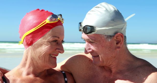 A senior Caucasian couple wearing swimming caps and goggles share a joyful moment on a sunny beach, with copy space. Their smiles and affectionate gaze reflect a sense of companionship and an active lifestyle.