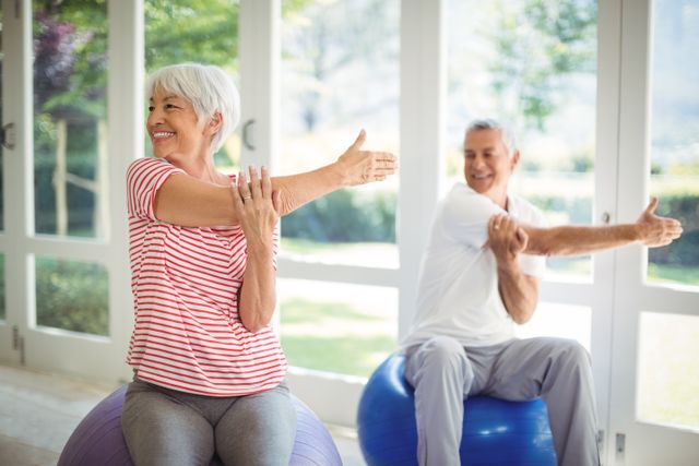 Senior couple smiling while performing stretching exercises on fitness balls in a bright indoor room. Ideal for promoting products or services related to senior health, home fitness routines, wellness programs, and physical therapy equipment.