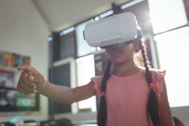 Young girl wearing virtual reality headset in classroom, pointing and interacting with virtual environment. Ideal for educational technology, modern learning methods, and innovative classroom experiences. Can be used in articles, blogs, and advertisements related to VR in education, child development, and future of learning.
