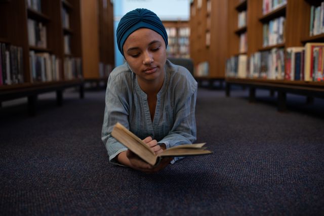 Front view of a biracial female student wearing a dark blue hijab studying in a library, lying on the floor, reading a book.