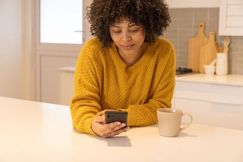 African american woman checking phone at kitchen counter wearing mustard sweater