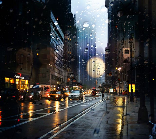 This is a rainy evening cityscape showing a busy downtown street with cars, pedestrians, and illuminated city lights reflecting on wet streets. Raindrops on a window provide an artistic effect. This image can be used for weather-related projects, urban lifestyle themes, or backgrounds for technology or transport-related content.