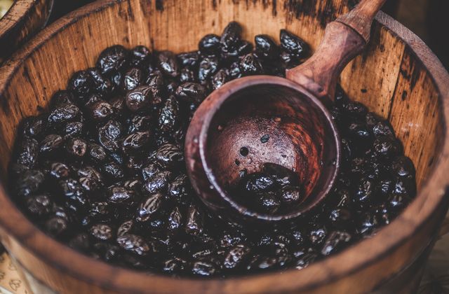 This image shows a wooden barrel filled with black olives, with a wooden scoop resting on top. Perfect for illustrating topics related to Mediterranean cuisine, traditional food preparation, healthy eating, organic farming, markets, and gourmet cooking. This can be used in marketing materials, food blogs, culinary websites, and health food promotions.