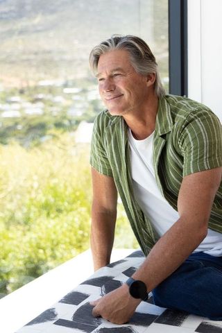 Senior man sitting on window bench, gazing at sunlit hills, wearing smartwatch, serene modern lifest