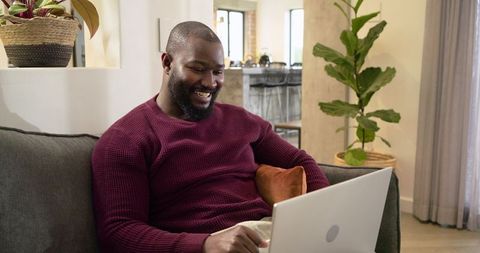 Mid-adult African American man smiling and typing on laptop while relaxing on living room couch