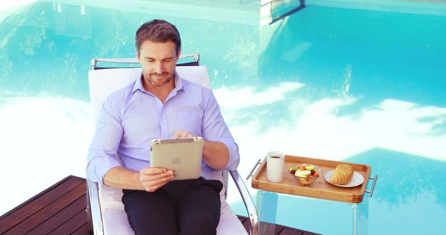 Businessman sitting by the pool, working on tablet while enjoying breakfast. Ideal for marketing materials for remote work, digital nomad lifestyle, productivity, summer retreats, and leisure activities.