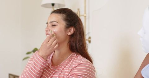 African american woman applying facial cream while friend wearing sheet mask at home