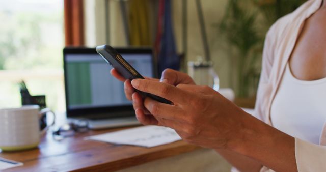 Hands using smartphone in home office. Ideal for illustrating themes about remote work, multitasking with technology, modern workspace, and productivity tips. Can be useful for articles or presentations on efficient work practices, tech usage in daily life, and maintaining productivity at home.