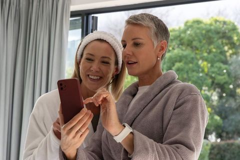 Mature woman and younger friend sharing smartphone while wearing cozy robes